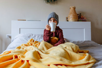 Girl wearing grey warm hat sits on the bed under warm blanket