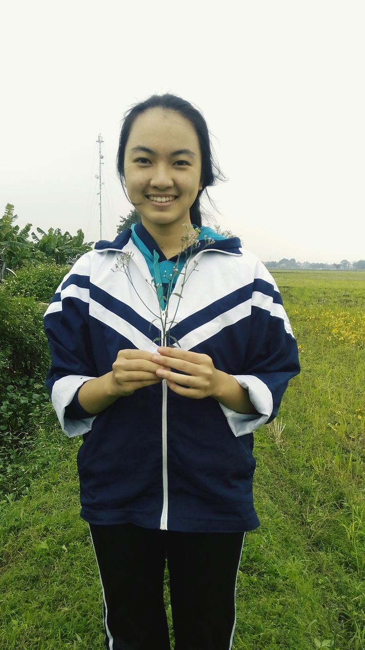 leisure activity, casual clothing, lifestyles, grass, three quarter length, standing, person, young adult, front view, looking at camera, field, green color, full length, portrait, clear sky, grassy, smiling, waist up