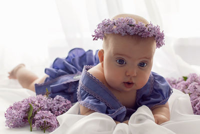 Chubby baby girl with big eyes in a purple dress lying on a white bed with lilac flowers 