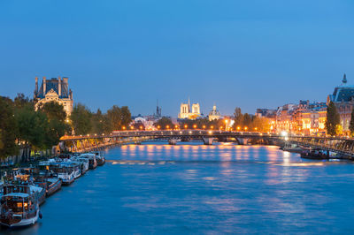 Illuminated buildings by river against clear blue sky