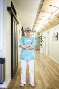 Full length portrait of mature female healthcare worker with arms crossed standing in hospital corridor