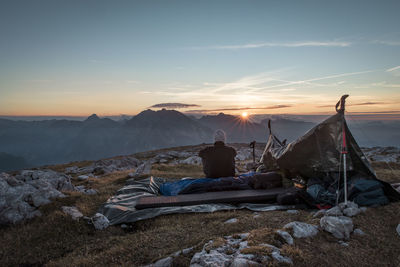 Rear view of man sitting by tent while looking at sunset over mountains against sky