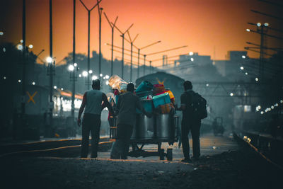 Rear view of people standing on street against sky during sunset