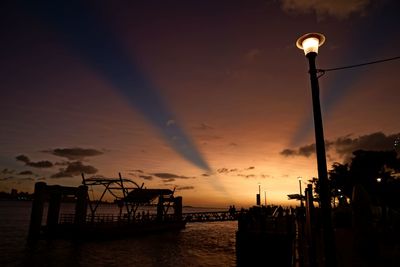 Silhouette street lights by sea against sky during sunset