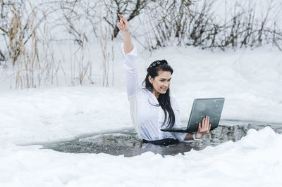 Portrait of smiling young woman using mobile phone while sitting on snow covered field