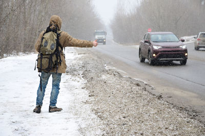 Rear view of hiker hitchhiking while standing at roadside during winter