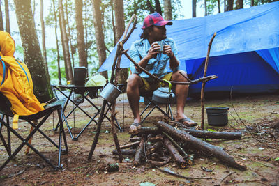 Man holding container while sitting on land by tent in forest