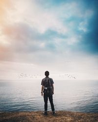 Rear view of man with camera standing by sea against cloudy sky during sunset