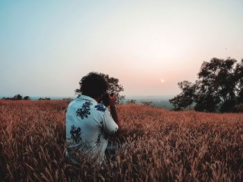 Side view of man photographing while sitting on land against clear sky during sunset