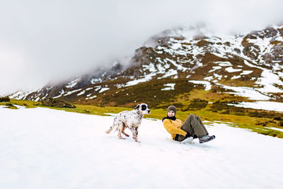 Happy woman sliding down slope covered with snow while happy english setter dog running aside in mountains