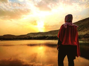 Rear view of man standing by lake against sky during sunset