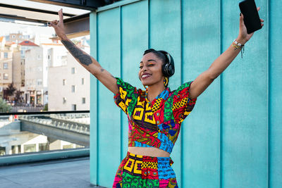 Cheerful ethnic woman in colorful clothes raising arms with smartphone and gesturing v sign while standing on terrace of modern building and listening to music in daytime