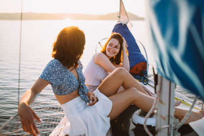 Side view of cheerful young female friends in stylish outfits smiling and talking while relaxing on sailboat against sunset sky during summer vacation