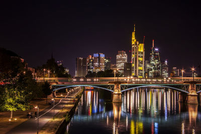 Illuminated bridge over river by buildings against sky at night