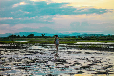 Rear view of girl standing on muddy field against cloudy sky during sunset