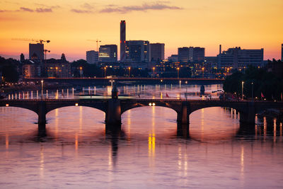 Bridge over river by illuminated buildings against sky at sunset