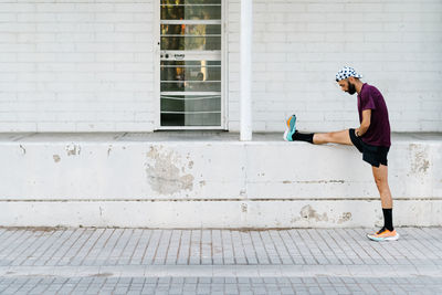 Side view of flexible male runner standing near stone fence on street and stretching legs before cardio workout in city