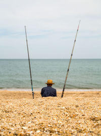 Man with fishing rods sitting at beach against sky