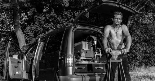 Portrait of shirtless man holding anvil on table against vehicle