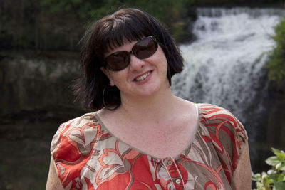 Portrait of smiling overweight woman wearing sunglasses against waterfall