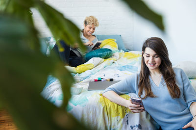 Smiling woman text messaging on smart phone while roommate lying on bed at home