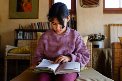 Front view of concentrated girl in lilac pullover reading a book in yellow armchair