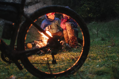 Cyclists arranging halt and warming at fireplace having warm beverage on a mug at green meadow at night