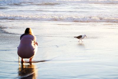 Woman crouching while looking at seagull in sea