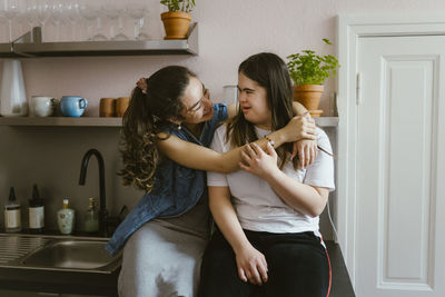 Smiling young woman hugging sister with down syndrome in kitchen at home