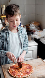 A caucasian boy in a shirt cuts pizza with salami with a round knife in the kitchen, on the table. 