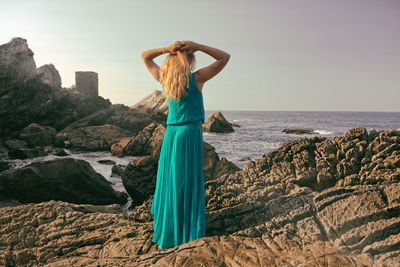 Rear view of mid adult woman with hands behind head standing on rock by sea against sky