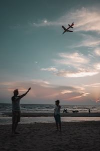 Mature man with boy flying kite on beach against sky during sunset