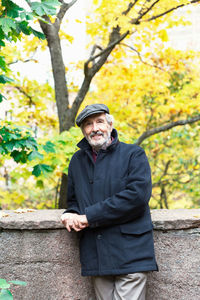 Portrait of confident senior man leaning on retaining wall while standing in park during autumn