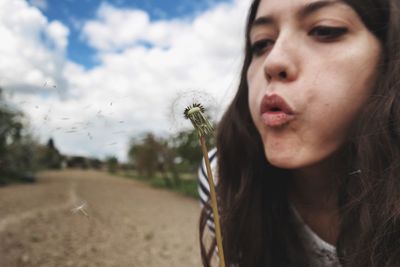 Close-up of young woman blowing dandelion seeds against sky