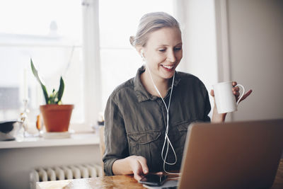 Happy woman listening to headphones while holding coffee cup at home