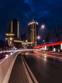 Light trails on road leading towards buildings against sky at night