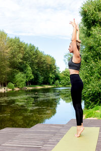 A woman stands on a green rug, by a pond , in summer, doing yoga.