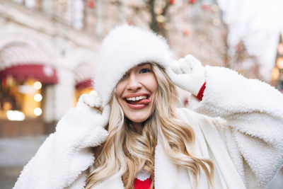 Young happy woman with curly hair in white fur hat having fun at the christmas fair winter street 