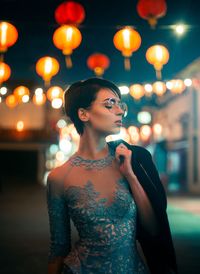 Young woman looking away while standing against illuminated lights at night