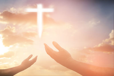 Low angle view of hands gesturing towards cross at sunset