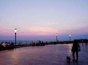 People walking on street by sea against sky during sunset