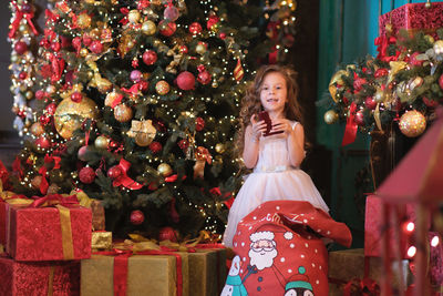 A little girl is taking out gift from new year's bag and smiling near the christmas tree background.