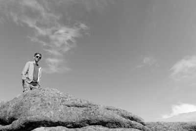 Low angle portrait of man in sunglasses standing on rock against sky