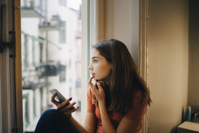 Thoughtful girl using phone while sitting at window sill