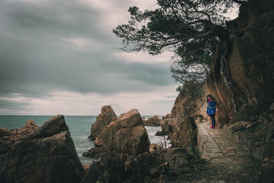 Woman standing on rock formation by sea against cloudy sky