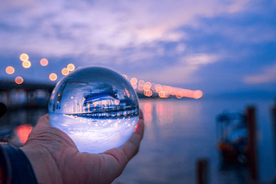 Cropped hand holding crystal ball by sea during sunset