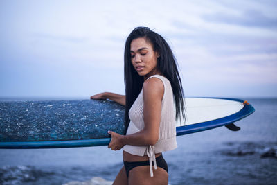Black multiracial woman lifestyle portrait by the ocean with surfboard