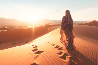 Rear view of woman standing on sand at beach against sky during sunset