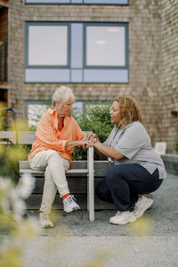 Full length of female nurse squatting while talking to retired senior woman sitting on bench at back yard