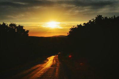 Silhouette trees by road against sky at sunset